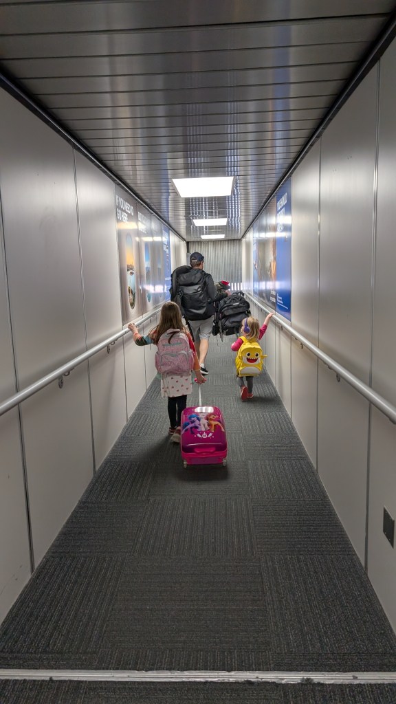 An adult male wearing a backpack walks down a jetway with two young children with backpacks following him